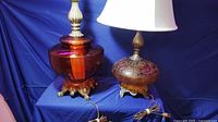 Photo showing two large orange pumpkin glass lamps on a blue cloth background. One lamp has a smooth orange glass base while the other features a crackle glass design. Both have brass necks and no shades.