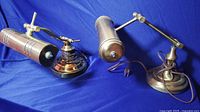 Photo showing two vintage brass desk lamps, placed side by side on a blue cloth background. One lamp has a rounded polished brass base and the other has a slightly different base and arm design.