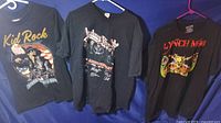 Front view of three black rock band t-shirts displayed on hangers against a blue backdrop showing Kid Rock, Judas Priest, and Lynch Mob designs.