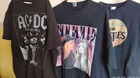 Front view of three rock band t-shirts hanging side by side: AC/DC, Stevie Nicks, The Beatles.