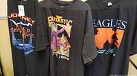 Three extra large black rock band T-shirts (Journey, Elton John, Eagles) displayed together showing graphic prints and size tags.