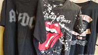 Three black band t-shirts hanging side by side featuring AC/DC, Rolling Stones, and ZZ Top logos and graphics.