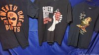 Three T-shirts hanging on a rod against blue fabric background: Twenty One Pilots, Green Day, and Smashing Pumpkins band shirts in black with various printed designs
