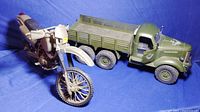 Photo showing angled front side view of the large green military transporter truck next to the beige motorcycle model, highlighting details on truck cab, front wheels, and bike body.