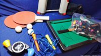 Mini pool table with bag of balls and cues, ping pong paddles and balls, jump rope on blue fabric background