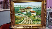 Frontal view of framed oil painting showing Italian village with trees and path under cloudy sky.