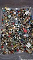 Top-down photo showing a large quantity of mixed small jewelry parts including beads, charms, and pendants in various colors and metal tones inside a transparent container.