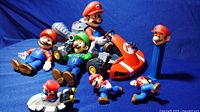 Super Mario Bros collection featuring large Mario and Luigi figures, Mario kart, small Mario figures, and a Mario head PEZ dispenser on blue background.