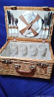 Open picnic basket showing cutlery with white handles, cloth napkins strapped inside the lid, and 4 clear glass goblets inside the lined basket interior