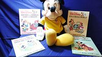 Full view of four vintage Walt Disney Mickey Mouse books arranged around a large plush Mickey Mouse talking doll in yellow outfit on blue background.