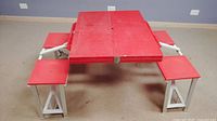 Red plastic fold-out picnic table fully set up showing tabletop and four attached seats with metal collapsible white legs.