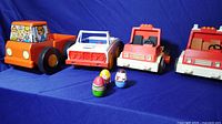 Four vintage toy trucks and three small Weeble Wobble figures displayed on a blue surface, showing fronts and side views.
