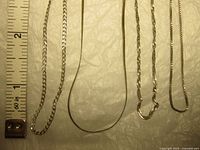 Top view of four silver chains arranged with measuring tape showing lengths ranging roughly from 7 to 18 inches. The chains have different link styles and thicknesses.