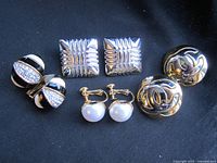 Front view of four pairs of earrings: black and white enamel clip-ons with rhinestones; silver-tone square clip-ons; gold-tone interlocking logo clip-ons; gold-tone faux pearl screw-back earrings