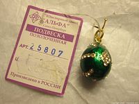 Photo showing close-up of green enamel egg pendant with gold-tone decorative motifs and tag with Russian text and article number 25807, indicating made in Russia.
