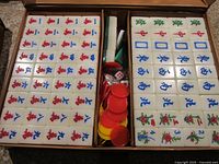 Open Mahjong set box showing two trays of Mahjong tiles with a compartment containing red and yellow chips, two dice, and other game accessories.