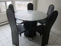 Photo showing round greenish marble-top dining table with cylindrical black pedestal base surrounded by five black tall-backed chairs on a white tiled floor in front of large window.