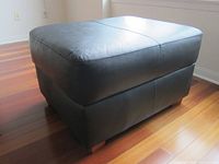 Black leather ottoman shown on a wood floor, highlighting the upholstery and wooden feet.