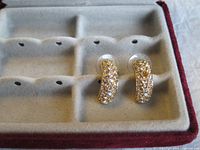 Pair of gold-tone Swarovski half hoop earrings with multiple clear crystals. One earring has one missing crystal, shown lying on velvet-lined earring holder.