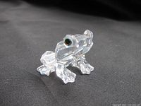 Close-up front angle of Swarovski clear crystal frog showing facets and black eyes, sitting on black fabric background.