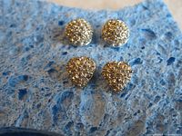 Top view of two pairs of stud earrings, heart-shaped gold tone and round silver tone with clear crystals, displayed on blue textured background.