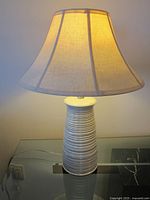 Photo of a 25-inch tall table lamp with a ribbed white ceramic base and beige fabric lampshade, turned on showing the warm light glow.