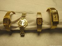 Four gold tone women's wristwatches arranged in a row showing front faces, including styles with mesh band, link band, expandable band, and cuff style band.