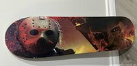Top view of skateboard deck showing Jason Voorhees' hockey mask and Freddy Krueger's face with artist signature on the bottom right corner
