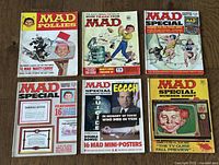 Top-down photo showing all 6 Mad Magazine issues laid out on wood floor, featuring bright colorful and illustrated vintage covers.