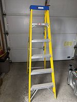 Yellow fiberglass step ladder with aluminum steps, blue top cap with Davidson logo, standing folded open in garage.