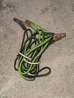 Photo of two used automotive booster cables with green/black and black insulation, metal clamps, coiled on the ground.