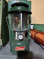 Front and side view of green Coleman 229 Series Two Mantle Lantern showing the glass globe, metal frame, and Coleman logo on the tank base. The lantern shows some wear and scratches.