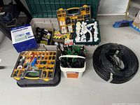 Full view of irrigation supply kit components including coiled hoses, digital timer, white connectors, black and green emitters, and assorted fittings organized in containers and toolboxes on concrete floor.