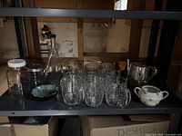 Shelf displaying the 9 clear glass mugs, green ceramic dish, silver metal teapot, white ceramic sugar bowl with floral pattern, and 3 glass jars including a bottle.