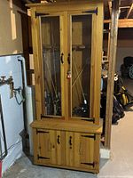 Tall wooden fishing rod cabinet with upper glass doors displaying fishing rods and lower cabinet with decorative hinges.