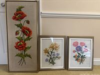 Group photo of three framed floral embroidery artworks leaning against a wall