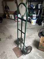 Full view of a green metal hand truck with a curved handle, pneumatic tires, and a rusted toe plate, standing on a concrete floor in a workshop area.