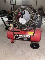 Full side view of red 8-gallon Husky air compressor with wheels and attached black coiled air hose.