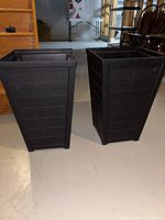 Two black tapered square planters shown side by side on a concrete floor in a warehouse or basement environment.