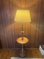 Wooden floor lamp with built-in small round table surface and pleated beige lampshade lit on wooden floor and paneled wall background.