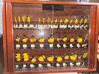 Front view of the Mastergrip router bit set inside a wooden case with glass sliding doors, showing three rows of yellow carbide tipped bits with labeled profiles.
