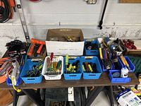 Wide view of table showing multiple hand tools including screwdrivers, brushes, and a propane torch arranged in baskets and boxes on a workbench.