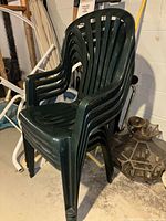 Four dark green stackable outdoor plastic chairs with armrests, stacked in corner against wall. Used condition visible.