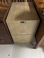 Gray metal two drawer filing cabinet placed between two wooden shelves or furniture pieces, photo taken to show the cabinet's condition and size.