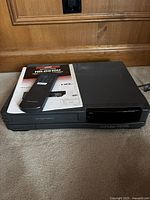Black JVC HR-D610U VHS player with remote and instruction manual on top on carpeted floor