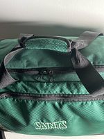Front view of the Adidas green and black polyester sport bag showing the padded handle, shoulder strap, and 'Saints' logo on the zippered front pocket.