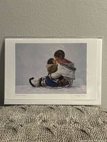 Full front view of the sealed art card showing the image of two children in winter clothes sitting and embracing on snow with a gray background.