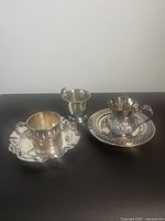 Photo showing two silver plated tea cups and plates, highlighting one cup with detailed filigree on handle and body and a smaller polished cup without handle.