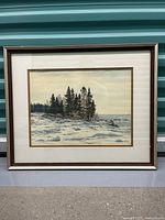 Full view of framed artwork showing snowy rocky shoreline with pine trees and water in background.