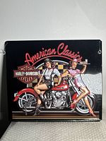 Full view of one embossed metal Harley-Davidson sign showing vintage red motorcycle and two pin-up girls with American Classics text and Harley-Davidson logo on black background.